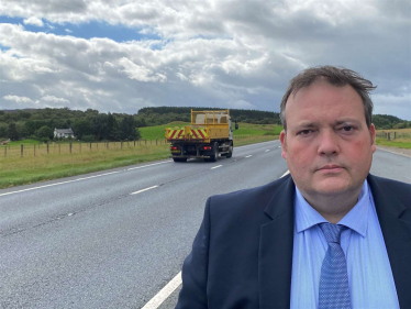 Jamie, wearing a suit, is standing beside the A9. A lorry is driving past.